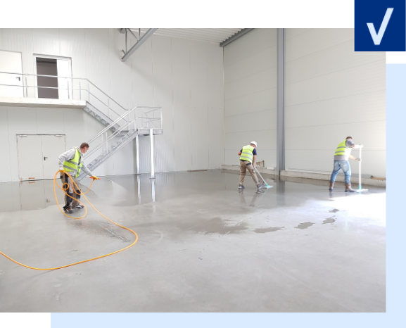 Construction Cleaning Three workers in safety vests clean a large, empty concrete floor inside an industrial building.