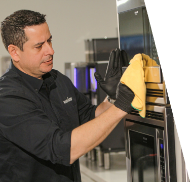 IMG_4274_EDIT-scaled-3 (1) Man wearing gloves cleaning a microwave oven with a yellow cloth in a kitchen setting.