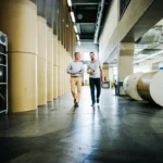 Two men walk and talk inside a large industrial facility with rolls of paper in the background.