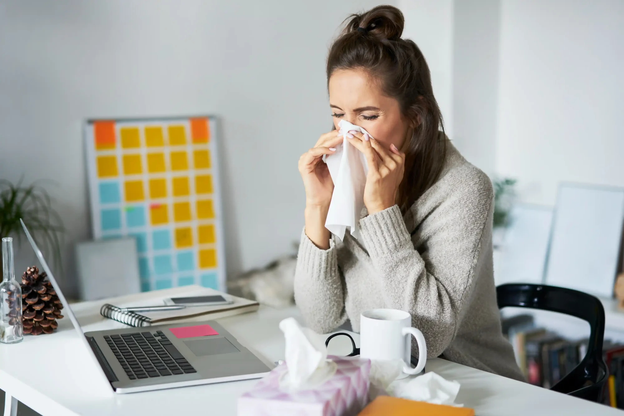 Woman sitting at a desk and blowing her nose with a tissue while working on a laptop.