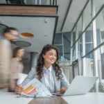 Woman working at a laptop with charts on the desk in a modern office as people walk by.