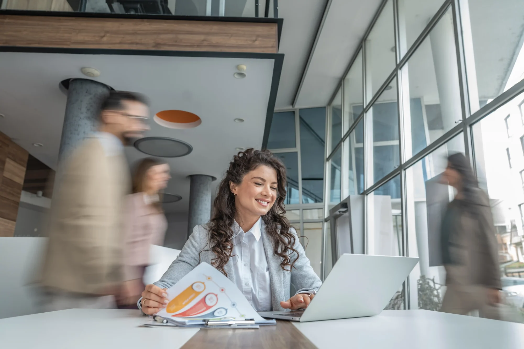 Woman working at a laptop with charts on the desk in a modern office as people walk by.