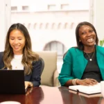 Two women sit at a conference table, one using a laptop and both smiling during a meeting.