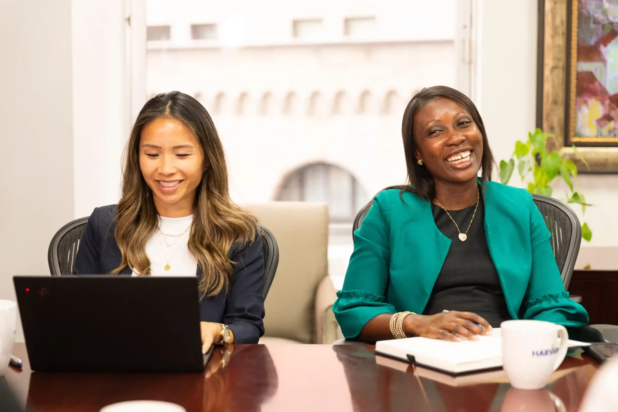 Two women sit at a conference table, one using a laptop and both smiling during a meeting.