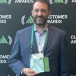 A bearded man in a suit holding an award in front of a branded backdrop.
