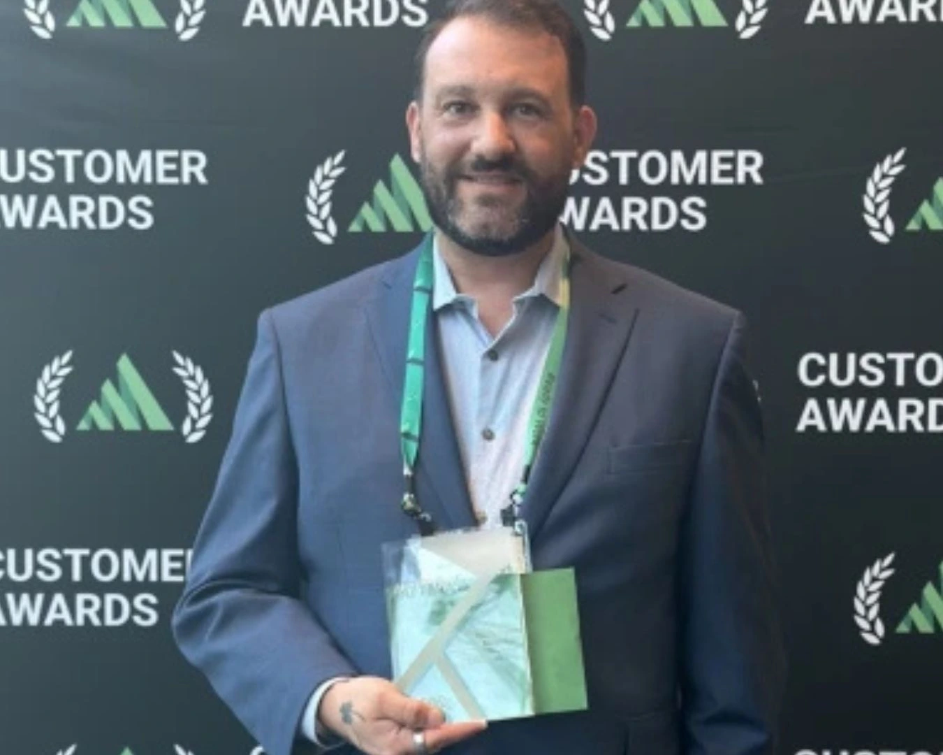 A bearded man in a suit holding an award in front of a branded backdrop.