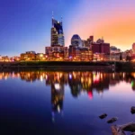 Nashville city skyline at sunset reflected in a calm river, with rocks in the foreground.