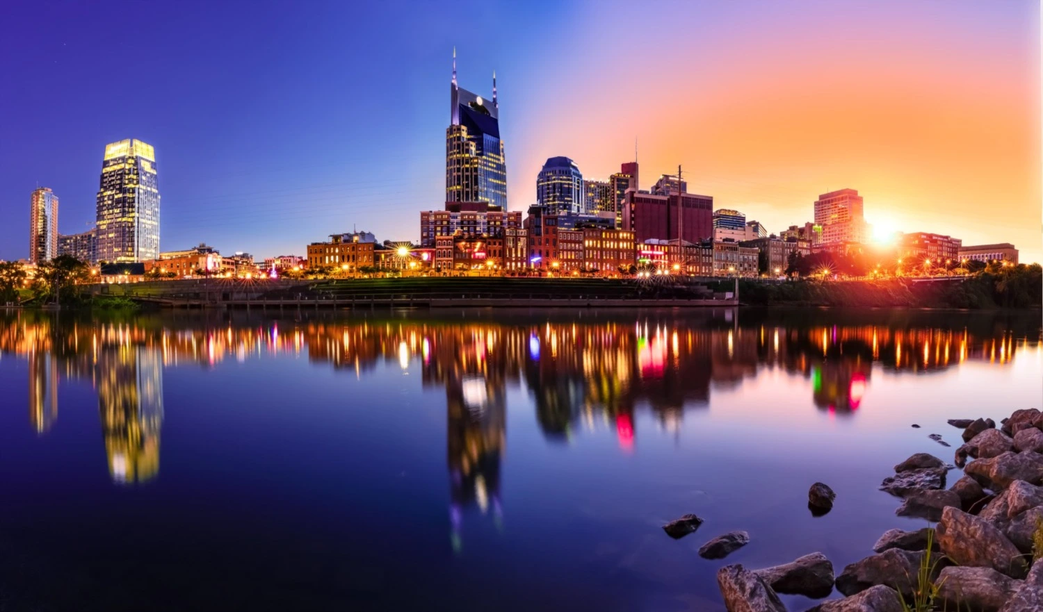 Nashville city skyline at sunset reflected in a calm river, with rocks in the foreground.