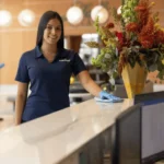 Woman in uniform cleans a lobby desk; large floral arrangement and computers are visible.