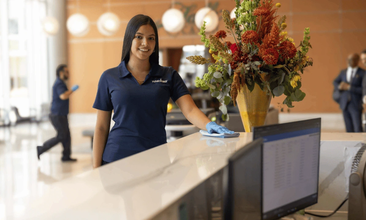 Woman in uniform cleans a lobby desk; large floral arrangement and computers are visible.